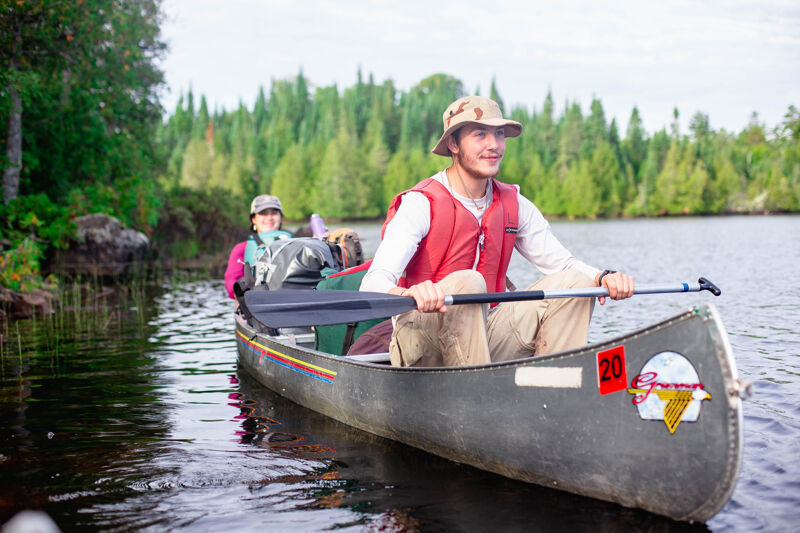 The image shows two people in a canoe on a lake. The person in the front is wearing a red vest and a hat, and is paddling the canoe. The person in the back is wearing a hat and a life jacket. The lake is surrounded by trees.